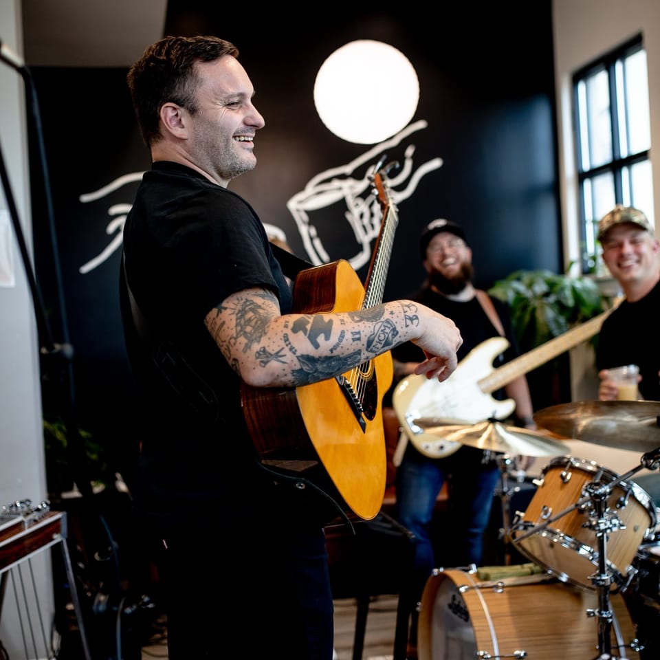 A smiling man with tattooed arms plays an acoustic guitar, with bandmates in the background.