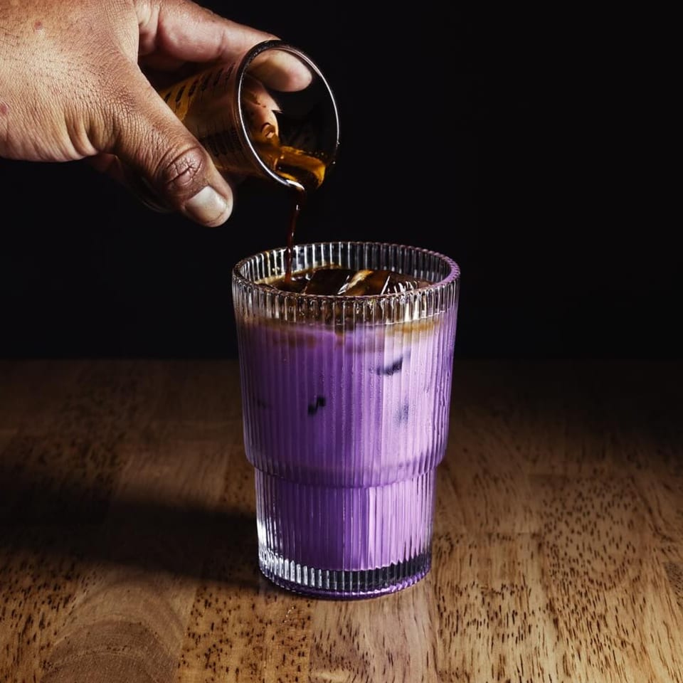 A hand pours dark espresso into a ribbed glass with purple iced milk on a wooden table.