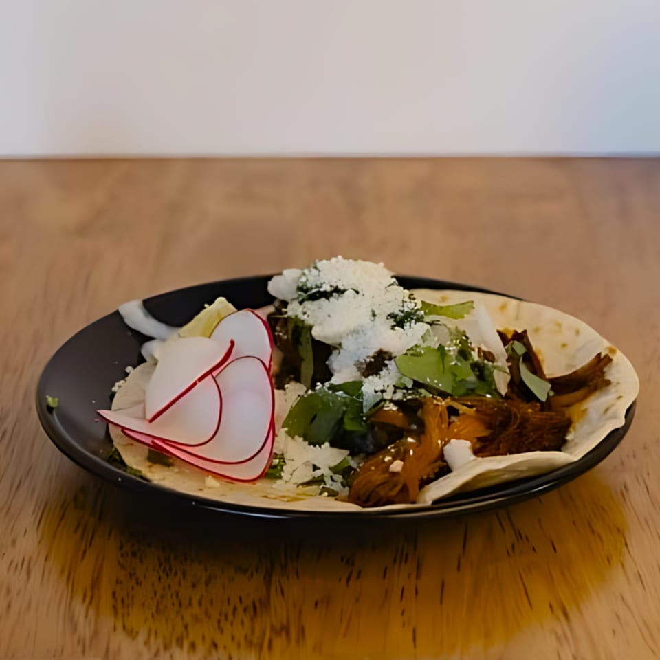 A black plate holds two tacos topped with meat, cilantro, cheese, and sliced radishes.