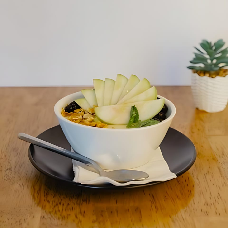 A white bowl of yogurt with sliced green apples, granola, and mint on a wooden table.
