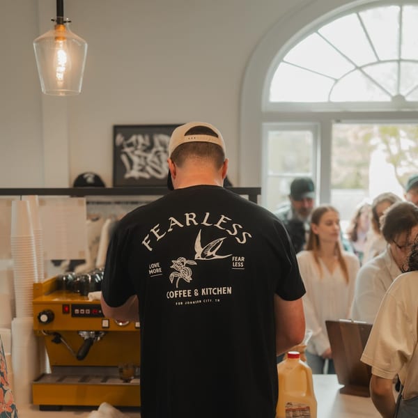 A barista in a Fearless Coffee & Kitchen shirt prepares drinks for customers at a cafe counter.