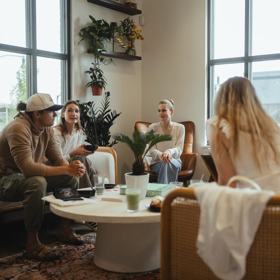 Four people conversing around a coffee table with drinks and plants in a bright cafe.