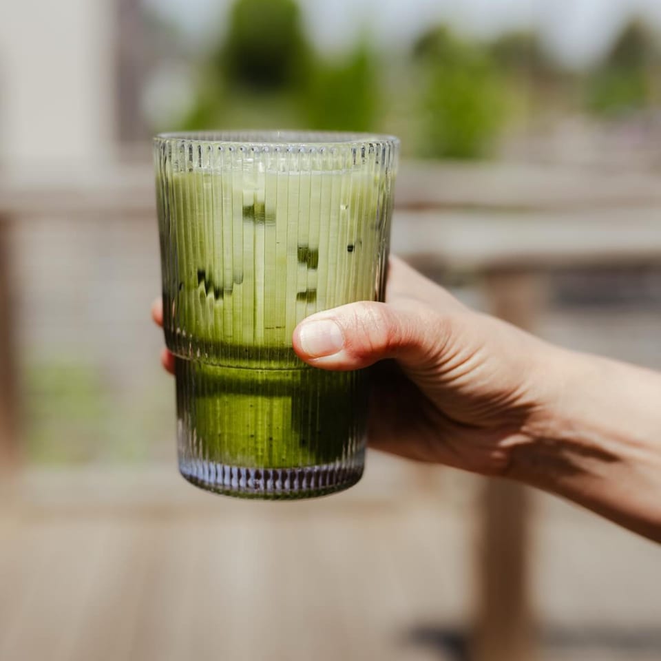 A hand holds a tall, ribbed glass filled with a vibrant green iced matcha latte.