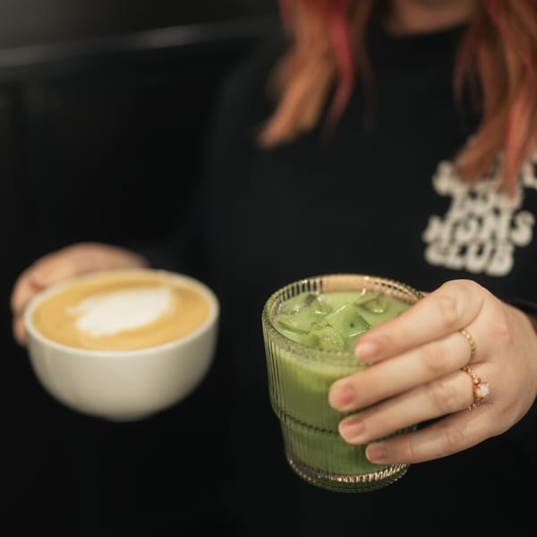 A person holds an iced matcha latte, with a hot latte in a white cup in the background.