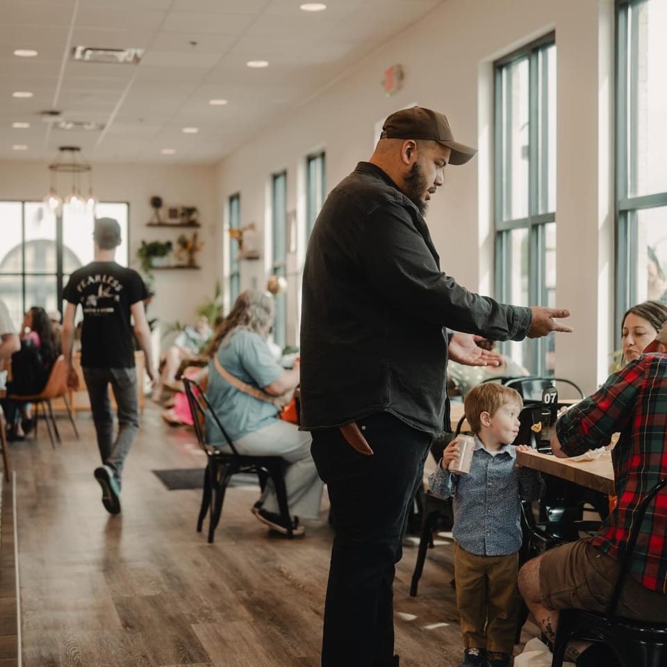 A man in a cafe talking to a family with a young child at a table.